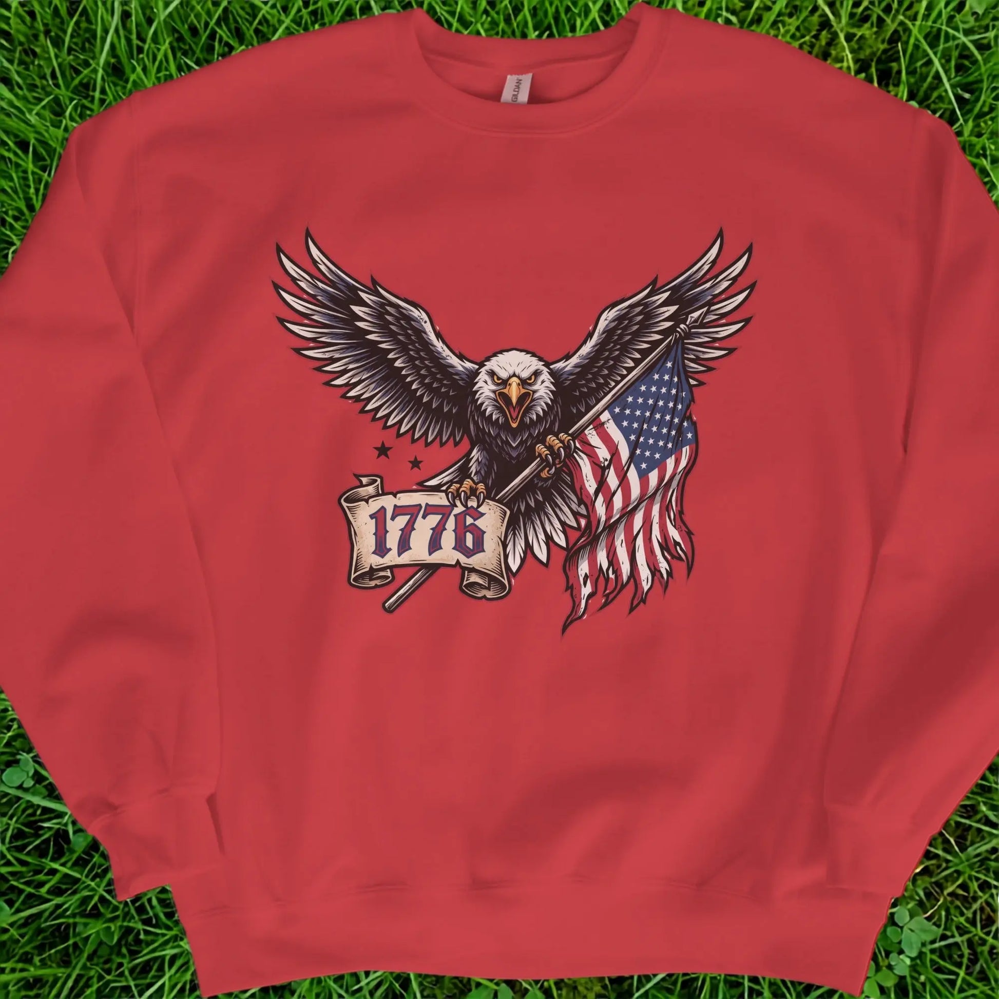 Bald eagle holding American flag and banner with "1776" text on red sweatshirt.