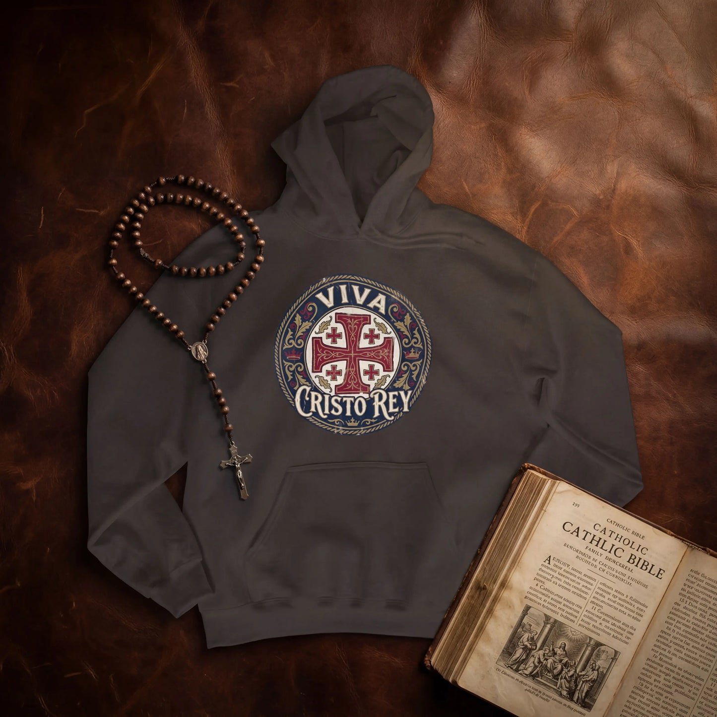 A dark gray hoodie laid flat on a brown leather surface, featuring a circular 'VIVA CRISTO REY' religious graphic on the chest. A wooden rosary with a cross and beads rests beside the hoodie, and an open vintage-style Bible is placed at the lower right.