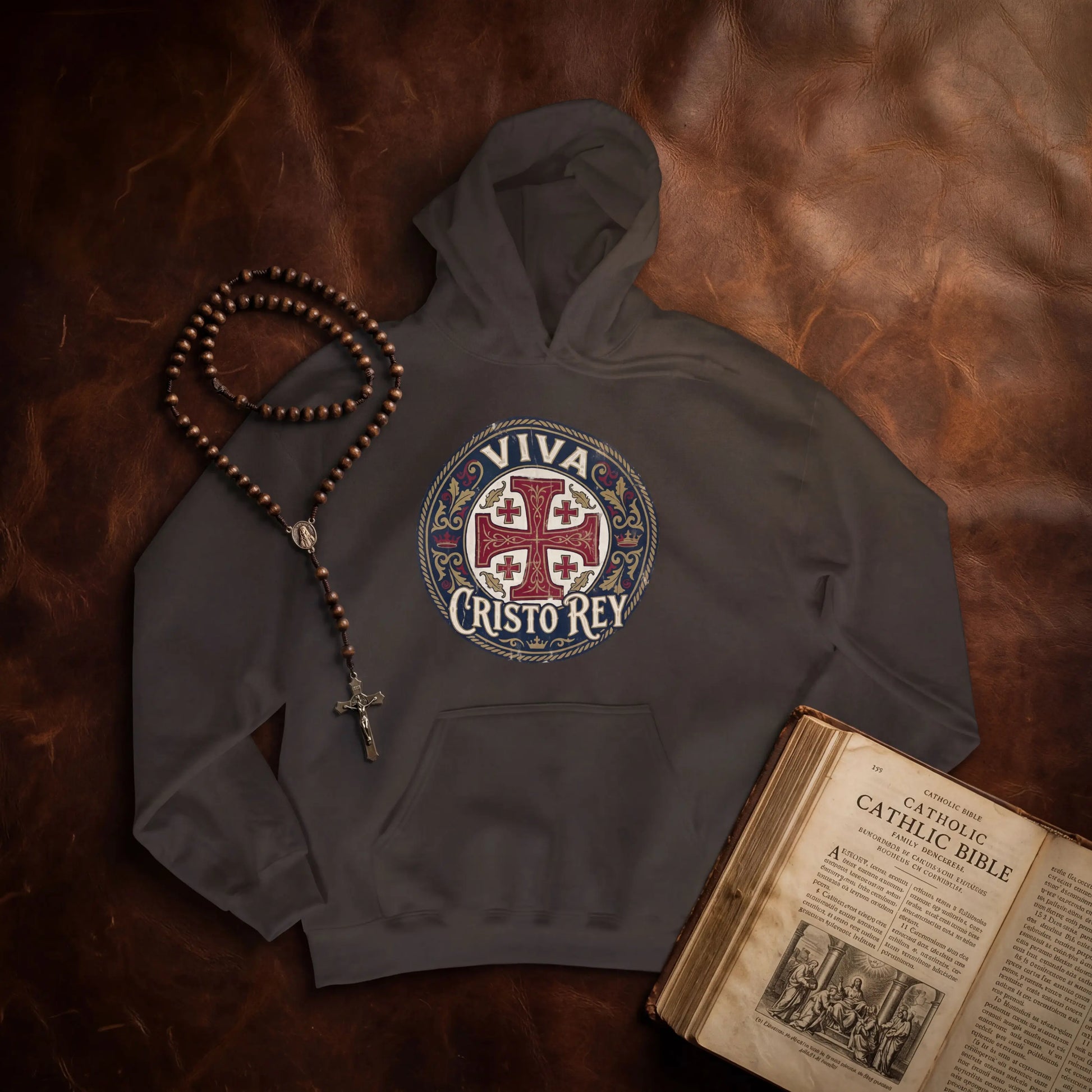 A dark gray hoodie laid flat on a brown leather surface, featuring a circular 'VIVA CRISTO REY' religious graphic on the chest. A wooden rosary with a cross and beads rests beside the hoodie, and an open vintage-style Bible is placed at the lower right.