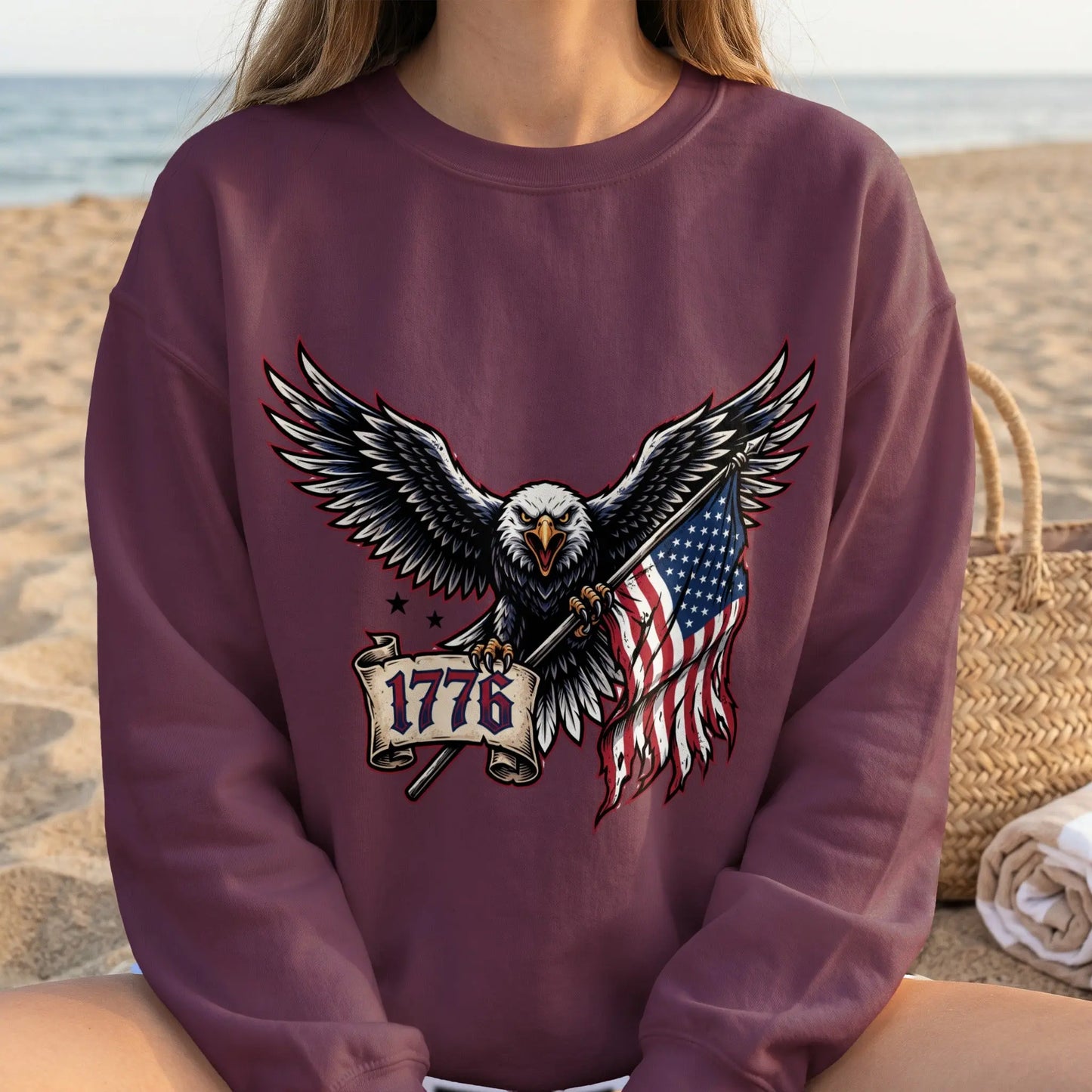 Bald eagle clutches US flag and banner with text "1776" on maroon sweatshirt.